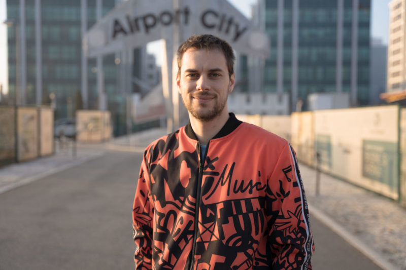 Ivan Kronokai, osnivač startapa Phobos, pozira ispred AirPort City-ja za kampanju Povezani. Ivan nosi narandžastu jaknu prošaranu crnim motivima.