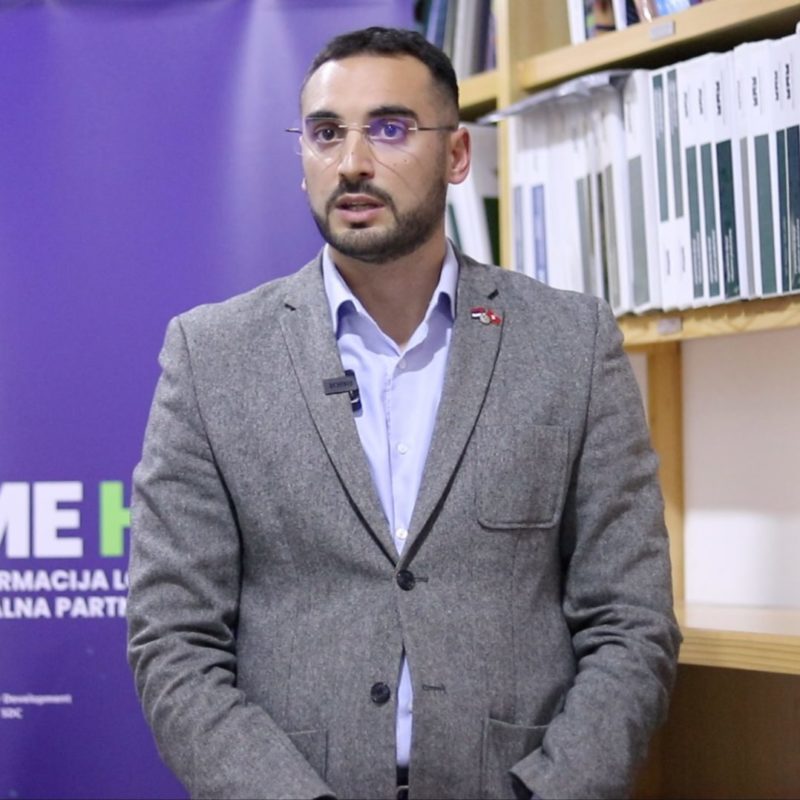 Aleksandar Goračinov, direktor SME HUB-a, stoji ispred plavo-ljubičastog banera sa logotipom istoimene inicijative. Aleksandar nosi svetlo plavu košulju i preko toga sivi sako.